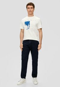 Katoenen t-shirt met ronde hals in off-white met een blauwe grafische opdruk; gecombineerd met marineblauwe broeken en witte sneakers. Eenvoudig ontwerp, casual pasvorm.