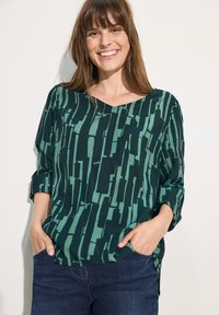 Top verde a fantasia con scollatura a V, tessuto textured e maniche a tre quarti, completato da una vestibilità ampia, abbinato a jeans scuri.