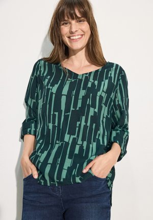 Top verde a fantasia con scollatura a V, tessuto textured e maniche a tre quarti, completato da una vestibilità ampia, abbinato a jeans scuri.