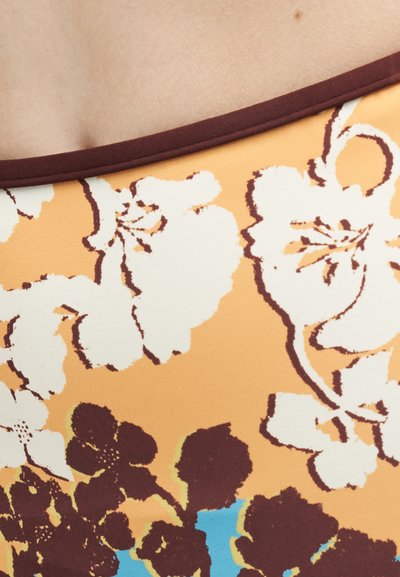 Κοντινή λήψη υφάσματος με πορτοκαλί φόντο, λευκά και σκούρα καφέ λουλουδένια σχέδια και σκούρο καφέ περίγραμμα κατά μήκος της πάνω άκρης.