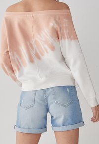 Mikina s designem na jednom rameni, světle růžová s bílým tie-dye vzorem; doplněná o roztrhané světle modré denimové šortky s ohrnutými lemy.
