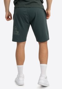 Mørkegrønne træningsshorts med elastisk talje, der har et trykt logo-detalje. Bæres med hvide sokker og sneakers.
