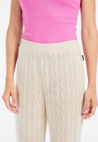 Beige gebreide broek met een ruitpatroon en subtiele uitsparingen, voorzien van een brede lichtgekleurde tailleband. Een roze ribbel- tank top is zichtbaar bovenaan.