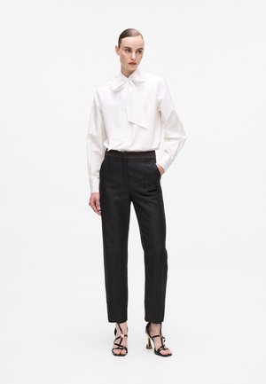 White long-sleeve shirt with a large bow tie, paired with textured black tailored trousers and strappy black high-heeled sandals.