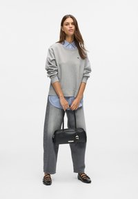 Γυναίκα που στέκεται, wearing γκρι oversized φούτερ πάνω από καρό πουκάμισο, φαρδύ γκρι τζιν, μαύρες επίπεδες γόβες, κρατώντας μικρή μαύρη τσάντα χειρός.