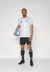 Mehrfarbiger Jersey in Pastellfarben mit Blau, Pink und Weiß; schwarze Shorts mit pinken Akzenten; schwarze Fußballschuhe und ein blauer Fußball.