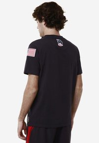 T-shirt nero a maniche corte con collo rotondo, caratterizzato da una piccola toppa con la bandiera americana sulla manica sinistra e logo sul colletto posteriore.