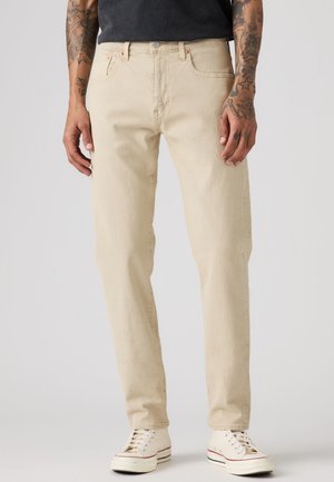 Homme portant un jean beige droit et des baskets blanches, avec des avant-bras tatoués et les mains détendues sur les côtés.