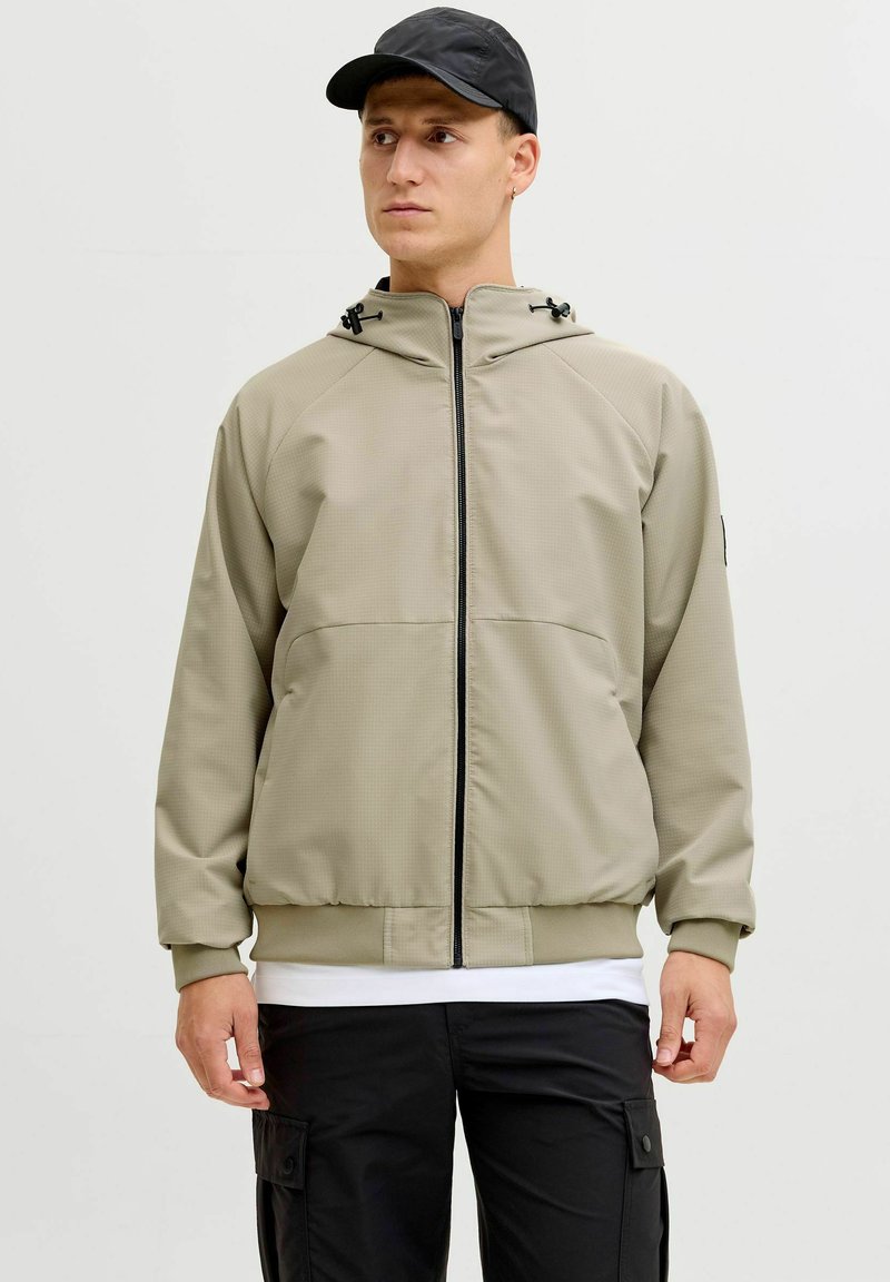 Homme portant une veste à capuche zippée beige, un pantalon cargo noir, une casquette noire, debout devant un fond clair uni.