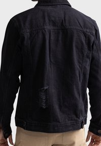 Chaqueta de mezclilla negra con cuello, costuras expresivas y una desgarradura desgastada en la parte baja de la espalda. Presenta puños abotonados y un dobladillo limpio.