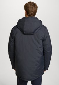 Mörkblå vadderad jacka med huva, som har en mjuk textur och en något glänsande yta. Designen inkluderar minimala detaljer och sömnadsdetaljer.