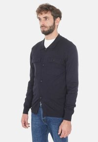 Uomo che indossa un cardigan scuro con bottoni sopra una camicia bianca e jeans blu, in piedi contro uno sfondo chiaro e uniforme, rivolto leggermente a destra.