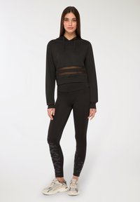 Giovane donna che indossa un hoodie nero corto con pannelli trasparenti e leggings neri con gambe inferiori a motivo e scarpe da ginnastica bianche.