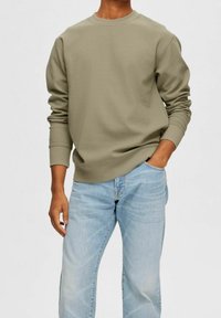 Olivově zelená mikina s kulatým výstřihem, žebrovanými manžetami a texturovanou látkou, doplněná světle modrými džíny. Klasický střih.