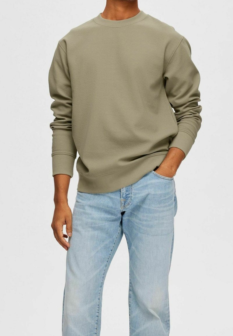 Olivově zelená mikina s kulatým výstřihem, žebrovanými manžetami a texturovanou látkou, doplněná světle modrými džíny. Klasický střih.