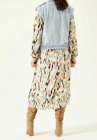 Jeansväst utan ärmar över en flerfärgad, mönstrad maxiklänning med långa ärmar; bruna knähöga stövlar fulländar looken.