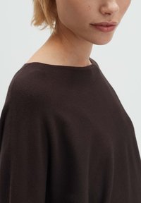Nahaufnahme einer Person mit kurzen blonden Haaren, die einen dunkelbraunen Strickpullover trägt. Zu sehen sind der Hals, die Lippen und der Schulterbereich.