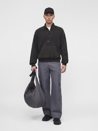 Homme portant une casquette noire, un sweat-shirt noir à fermeture éclair au quart, un pantalon cargo gris et des chaussures noires, tenant un grand sac gris dans une main.