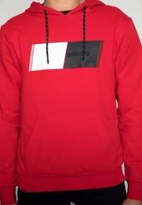 Felpa rossa con design rettangolare nero e bianco sul petto, che presenta un logo. Dettagli con cordini neri e tasca a marsupio frontale.
