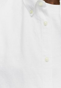 Witte linnen overhemd met een button-down kraag, voorzien van lichtgekleurde knopen en een textured stof, gedeeltelijk zichtbaar op de afbeelding.