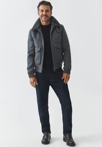Bomber jacket grigio in misto lana con colletto in pelliccia sintetica, due tasche anteriori a toppa e polsini a costine, indossato sopra un maglione scuro e jeans.