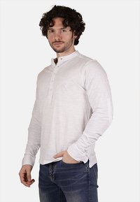 Camicia henley bianca a maniche lunghe con una sottile texture, caratterizzata da un colletto arrotondato, placket con tre bottoni e polsini aderenti. Abbinata a jeans scuri.
