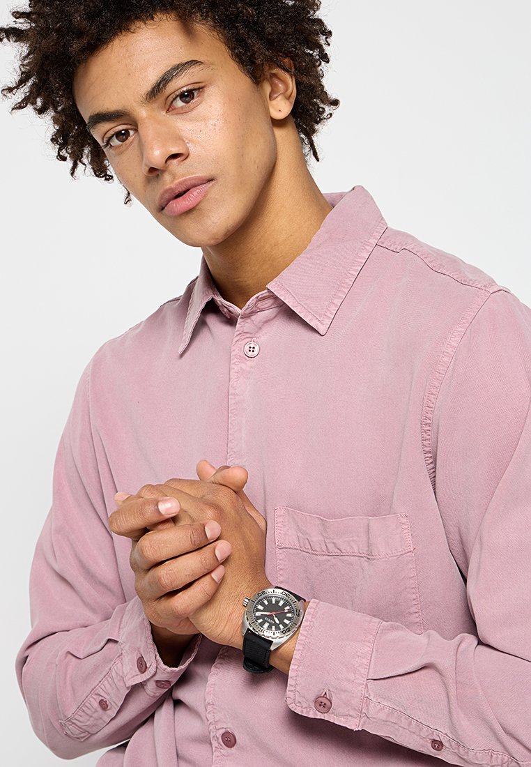 Camisa de botones de color rosa claro en un corte relajado, con un bolsillo en el pecho y una textura distintiva, combinada con un reloj plateado y correa negra.