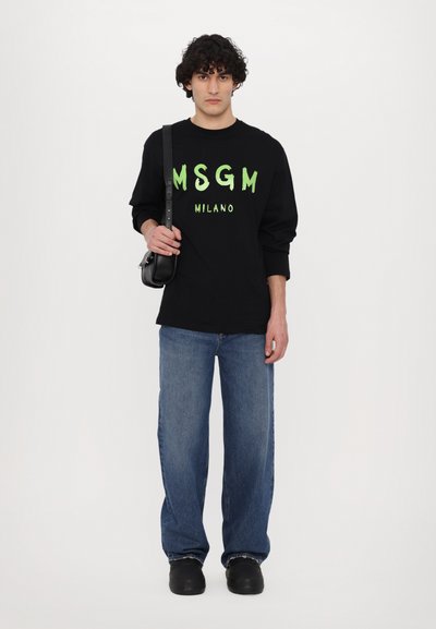 Svart langermet T-skjorte med grønn "MSGM MILANO" tekst, kombinert med løstsittende blå jeans og svarte slip-on-sko. Enkel skulderveske inkludert.