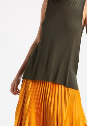 Ärmelloses olivgrünes Oberteil kombiniert mit einem leuchtend orangefarbenen plissierten Rock, gezeigt vom Oberkörper bis zu den Hüften vor schlichten Hintergrund.