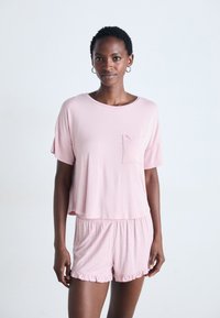 Kvinna som står i en rosa kortärmad topp och matchande ruffle shorts med ett vitt prickmönster mot en enhetlig bakgrund.