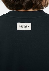 Schwarzer Pullover mit geripptem Kragen und einem weißen Stoffetikett auf der Rückseite, das "SENSES .THE LABEL" in fetten schwarzen Buchstaben trägt.