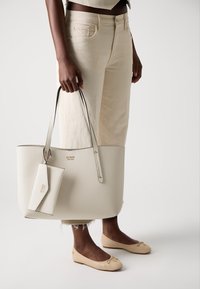 Sac fourre-tout blanc texturé avec des poignées grises et une étiquette assortie, accompagné de ballerines beiges avec un accent doré. Design minimaliste, style décontracté.