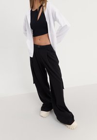 Pantalon cargo noir à coupe décontractée, avec poches latérales, ceinture, et un sweat à capuche zippé léger blanc porté par-dessus un crop top noir.