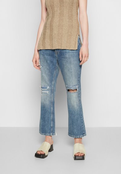 Top de malha canelada bege, jeans retos azuis desgastados com bainhas desfiadas, e mules em creme de biqueira aberta com salto bloco.