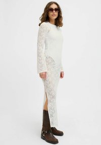 Witte kanten jurk met lange mouwen en zijsplitten, gedragen met bruine enkellaarsjes met gespen. Eenvoudige achtergrond, model staat iets naar rechts gekeerd.