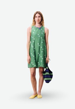 Jeune femme portant une robe verte sans manches à pois blancs, des ballerines jaunes, tenant un sac rayé bleu et vert, debout face à nous.