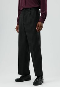 Zwarte broek met krijtstrepen en rechte pijpen, voorzien van zijzakken en een tailleband. Gecombineerd met zwarte loafers en een bordeauxrode blouse met knopen.