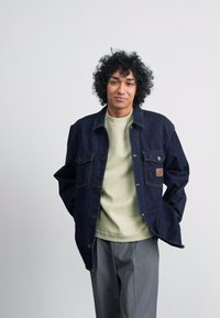 Dark denim jacket with front pockets, paired with a light green shirt. Gray trousers complete the outfit. Model stands with hands in pockets.