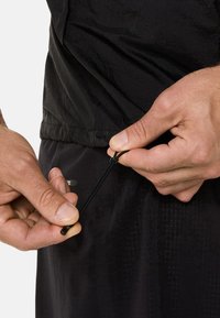 Sort letvægtsbeklædning med en struktureret overflade, der har et justerbart snor håndteret i den ene hånd. Nærbillede af hænder, der griber fat om snoren.