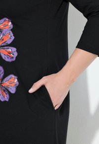 Svart klänning med blommönster i lila och orange på vänster sida, utrustad med en sidficka. Mjuk, lättviktsmaterial med avslappnad passform.