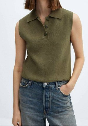 Kobieta nosząca bezrękawnik oliwkowy zielony dzianinowy top polo z trzema guzikami, w połączeniu z wysokim stanem niebieskich dżinsów, jedna ręka w kieszeni.