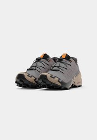 Trailrunning-Schuhe in Grau mit strukturierten Oberflächen. Ausgestattet mit einem orangefarbenen Ziehband und schwarzen Gummisohlen mit tiefen Stollen für optimalen Halt.