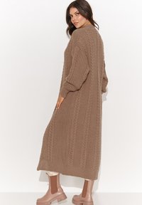 Długi brązowy kardigan dzianinowy z teksturowanym ribbingiem i luźnym krojem. Połączony z jasnoróżowymi botkami za kostkę dla kontrastu.