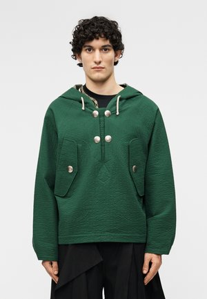 Νέος με κατσαρά μαλλιά που φοράει μια υφασμάτινη πράσινη κουκούλα με ασημένια κουμπιά και μεγάλες μπροστινές τσέπες, στέκεται μπροστά σε απλό φόντο.