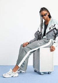 Tuta grigia e bianca con giacca cropped, caratterizzata da lettere in grassetto e righe. Il modello indossa occhiali da sole neri e sneakers bianche chunky.