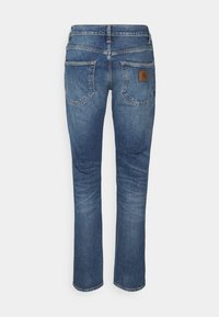 Jeans de mezclilla azul con corte recto, cinco bolsillos y una parche de cuero marrón en la parte posterior de la cintura. Desgaste ligero y costuras visibles.