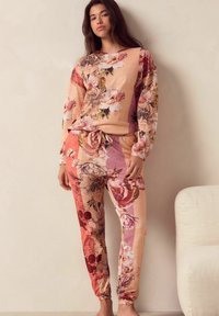 Set de loungewear florale dintr-un material moale, cu o paletă de culori crem și roz, având modele florale variate. Stilul include un hanorac și pantaloni jogger.