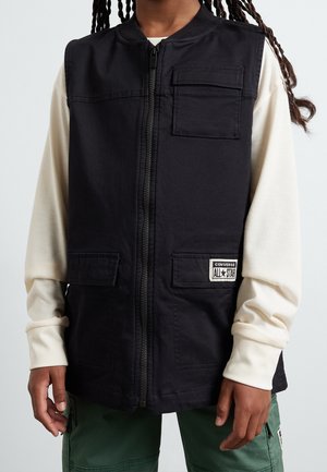 Sort vest med lynlås og forlommer med klap båret over en langærmet cremefarvet skjorte og grønne cargobukser, der viser del af torso og hænder.