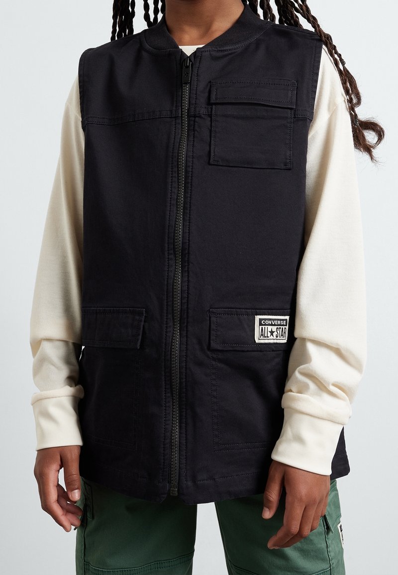 Gilet noir zippé avec poches à rabat devant, porté sur une chemise crème à manches longues et un pantalon cargo vert, montrant une partie du torse et des mains.