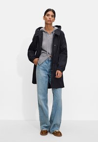 Czarny płaszcz przeciwdeszczowy z kapturem, noszony na szarym rozpinanym swetrze, w zestawie z jasnoniebieskimi dżinsami i butami wsuwanymi w panterkę.